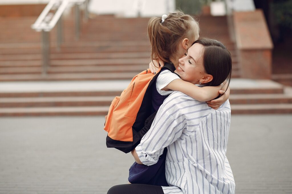 Uma mãe abaixada, abraçando afetuosamente sua filha. A criança possui uma mochila nas costas.